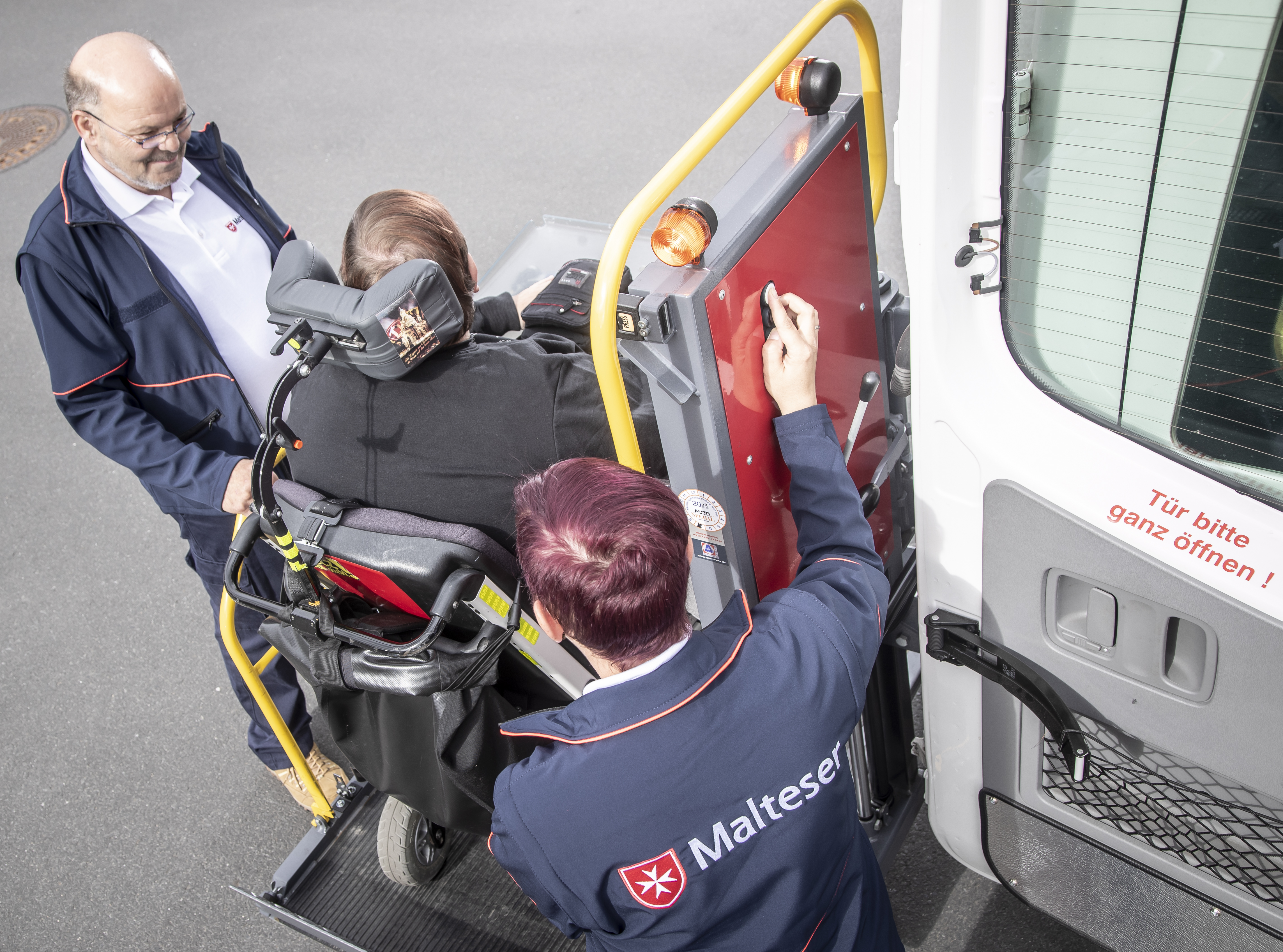 Zwei Mitarbeiter der Malteser heben mit einer Hebebühne eine Person im Rollstuhl in ein Fahrzeug